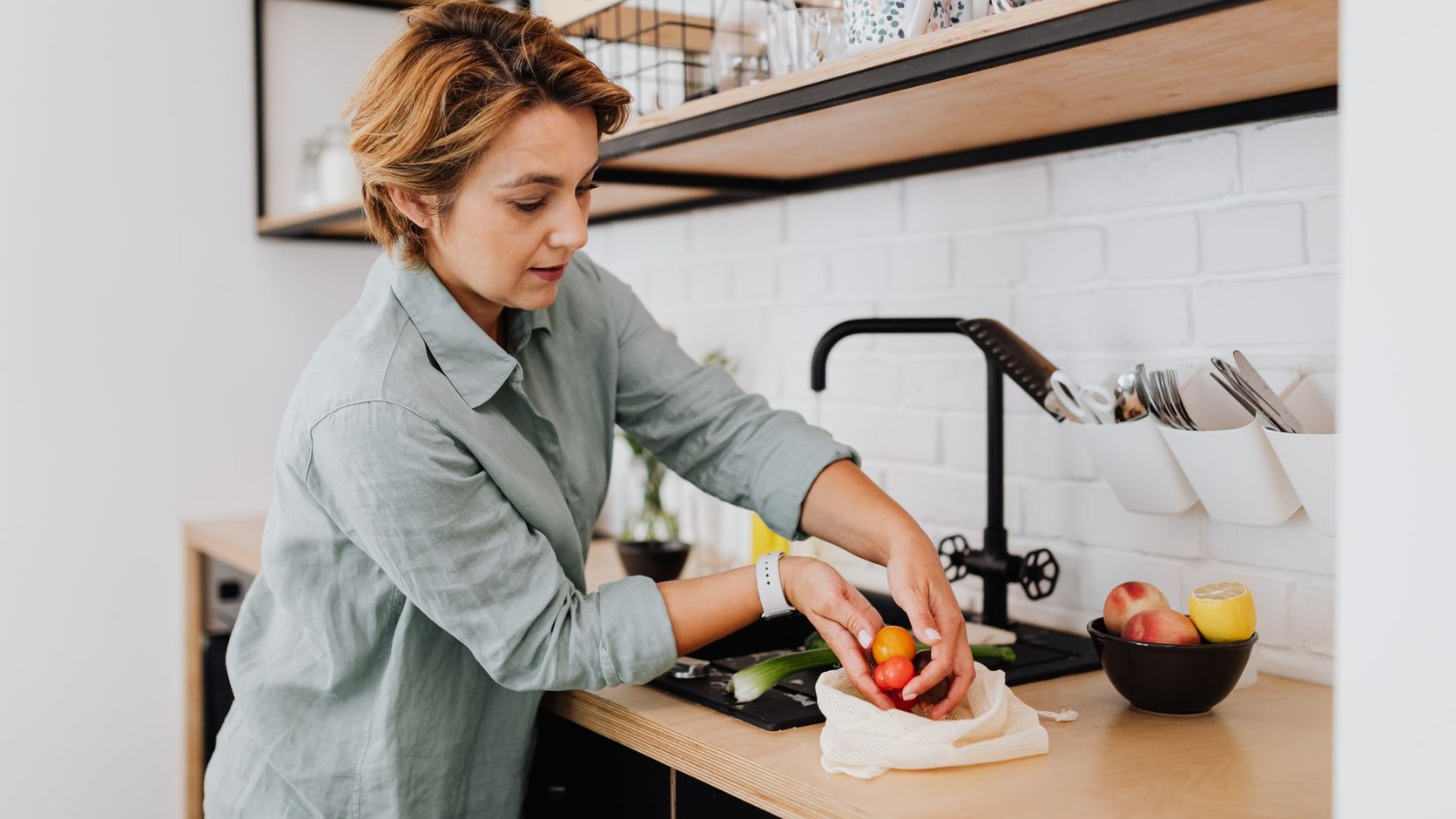 Mulher adulta organizando frutas e legumes em cozinha de casa com luz natural