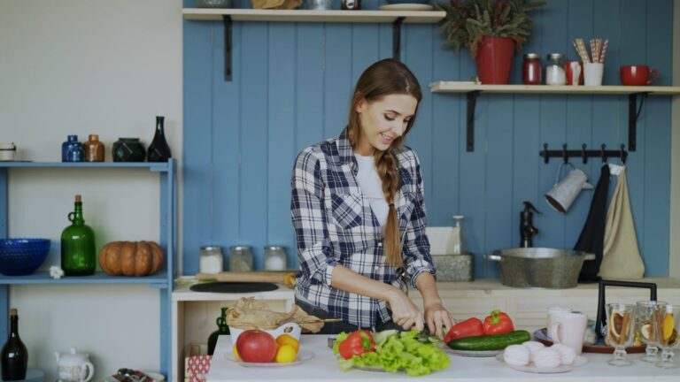 Mulher jovem picando legumes em uma cozinha caseira, com ingredientes frescos na bancada.